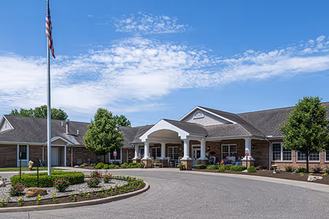 Front exterior view of Charlotte Stephenson Manor, a single-story brick building with a covered entrance supported by white columns. There is a circular driveway with landscaped flower beds and an American flag on a flagpole. The sky is blue with scattered clouds.