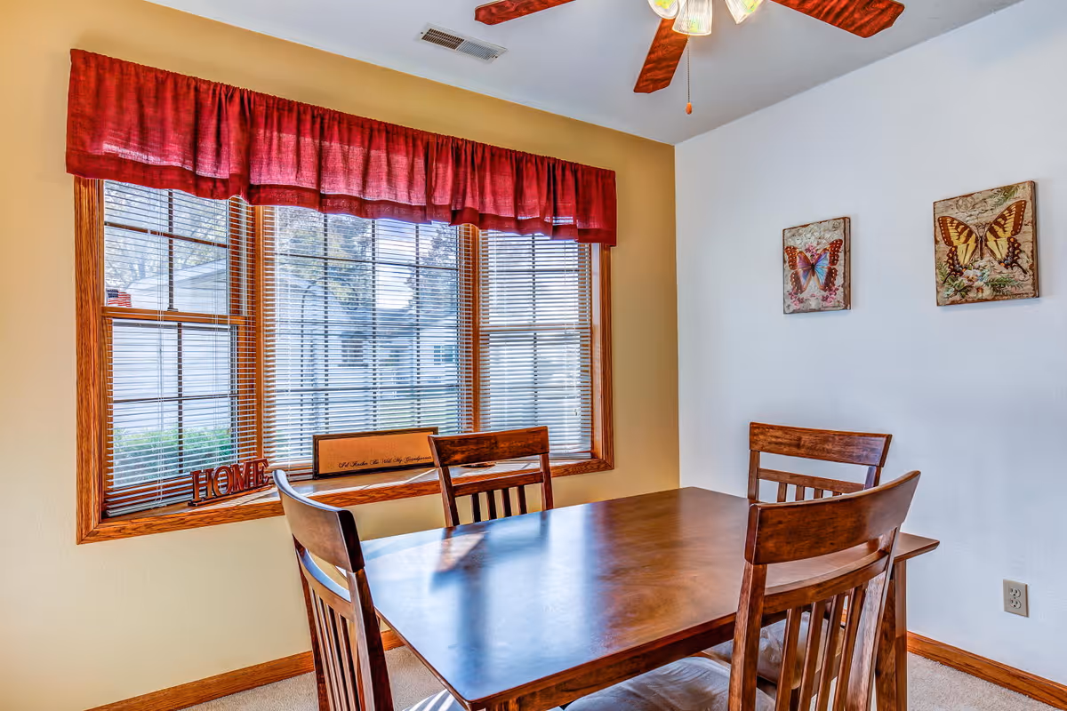 A dining area with a wooden table and four matching chairs. The room has a large window with wooden blinds and a red valance. On the windowsill, there is a decorative sign that says 'HOME' and another rectangular sign. Two butterfly-themed wall decorations hang on the white wall. The ceiling has a fan with wooden blades and light fixtures.