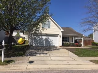 Single-story house with an attached two-car garage, driveway, front lawn, tree, and mailbox under a clear blue sky.