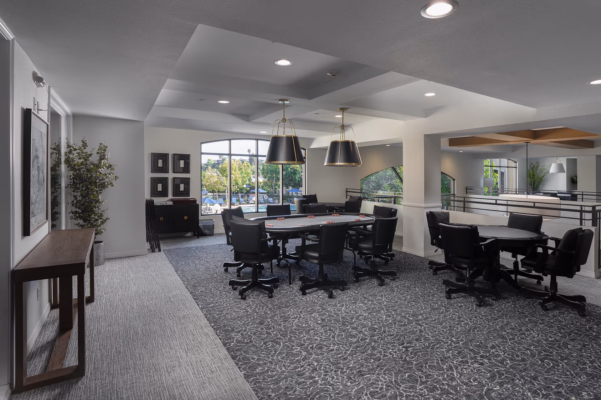 Interior view of a modern common area in Woodland Village featuring two round tables with black swivel chairs on a patterned carpet. Large windows allow natural light to brighten the space, and two large pendant lights hang from the ceiling. There is a console table with a plant and framed artwork on the left side of the room.