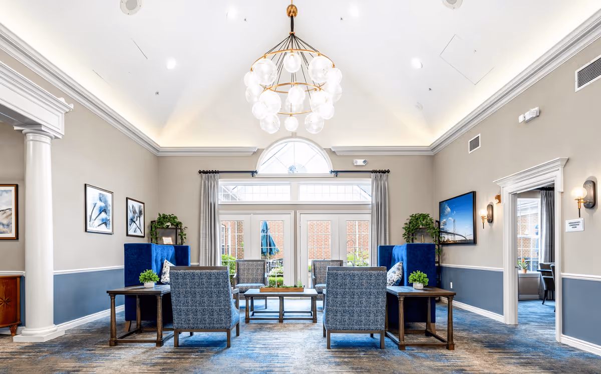 A bright and spacious living room area in Waltonwood Twelve Oaks featuring high vaulted ceilings with a modern chandelier, large windows with curtains allowing natural light, blue and gray upholstered chairs arranged around wooden tables, decorative plants, framed artwork on the walls, and an open doorway leading to another room.