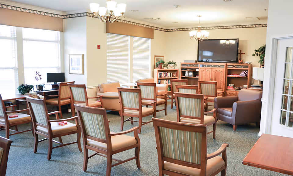 A well-lit common room with multiple wooden chairs arranged in rows facing a large flat-screen TV mounted on a wooden entertainment center. The room has beige walls, large windows with blinds, a couple of sofas, a brown armchair, and a small desk with a computer in the corner. There are plants and framed pictures decorating the space.