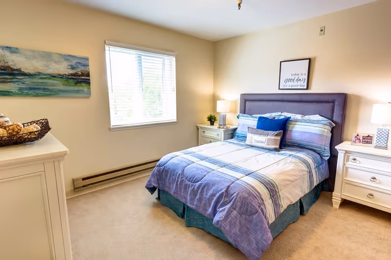 A bright and cozy bedroom with a neatly made bed featuring a purple and blue striped comforter and multiple pillows. The bed has a padded headboard and is flanked by two white nightstands, each with a lamp. A framed quote hangs above the bed, and a window with white blinds lets in natural light. There is a white dresser with a decorative basket on top and a landscape painting on the wall.