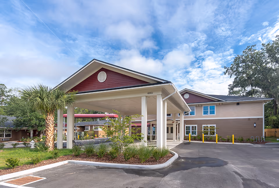 Covered entrance drive-through and front facade of a two-story senior living building with landscaping and a palm tree under a blue sky.