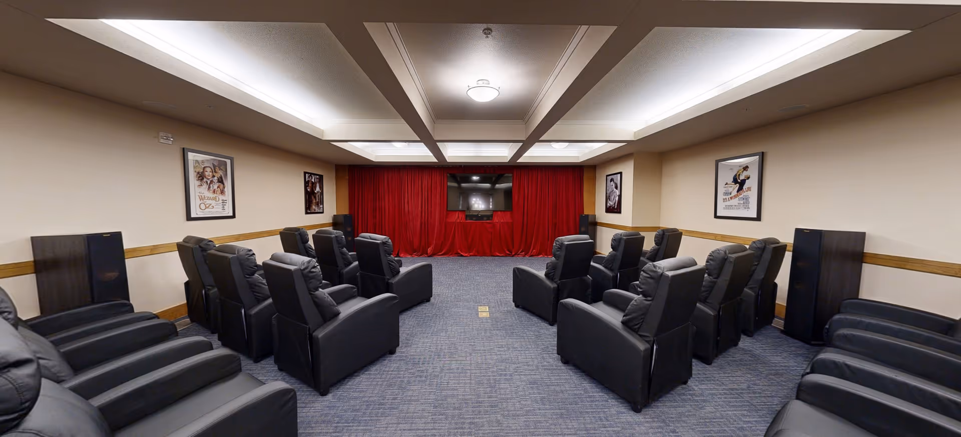 A small theater room with two rows of black recliner chairs facing a large flat screen TV mounted on a wall with red curtains. The room has beige walls with framed movie posters and a blue carpeted floor.