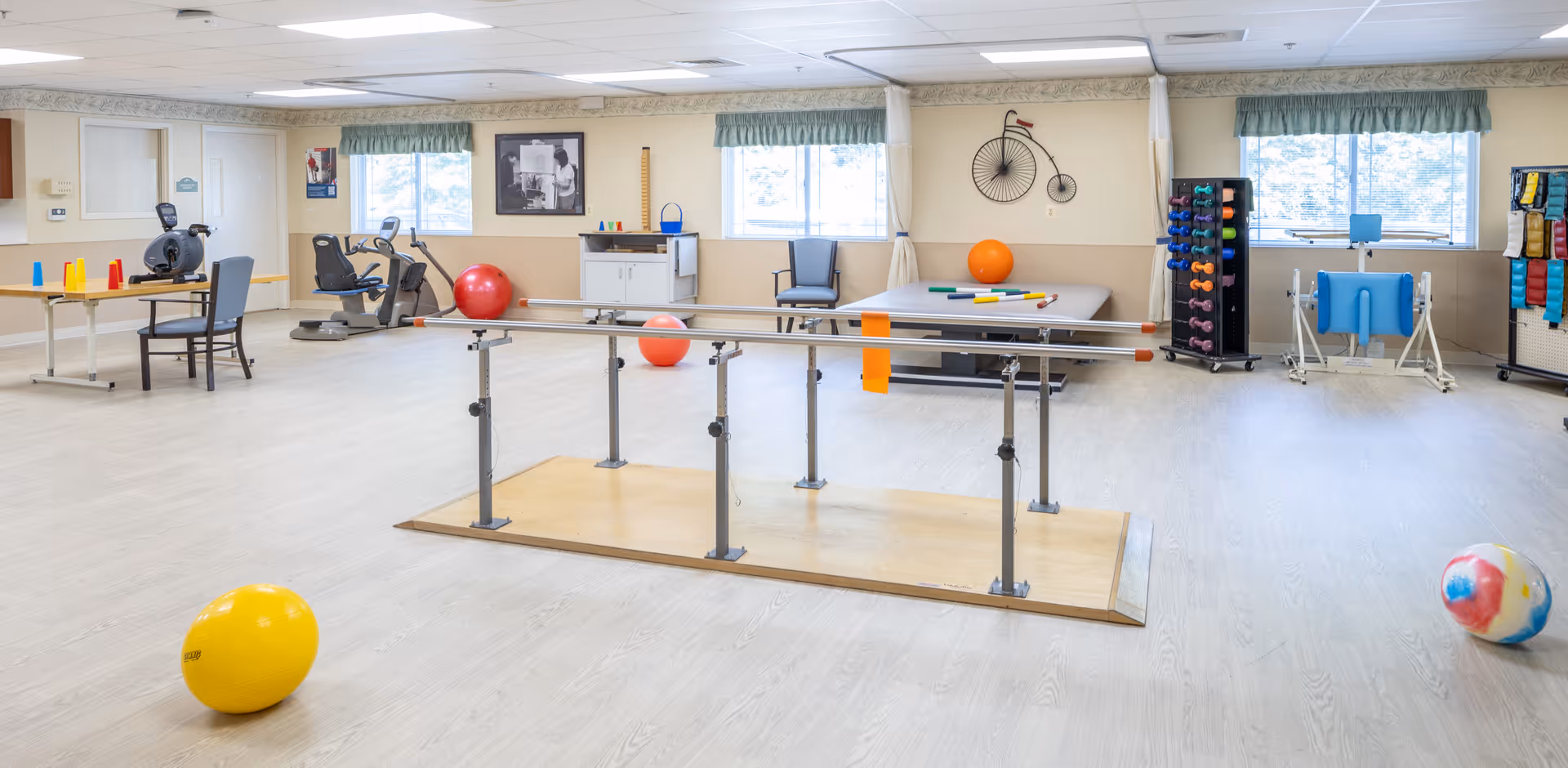 A spacious physical therapy or rehabilitation room with parallel bars in the center for walking exercises. The room contains exercise equipment including stationary bikes, therapy balls, dumbbells on a rack, and various therapy tools. Large windows with green valances let in natural light, and the walls are decorated with a vintage bicycle picture and a black and white photo.