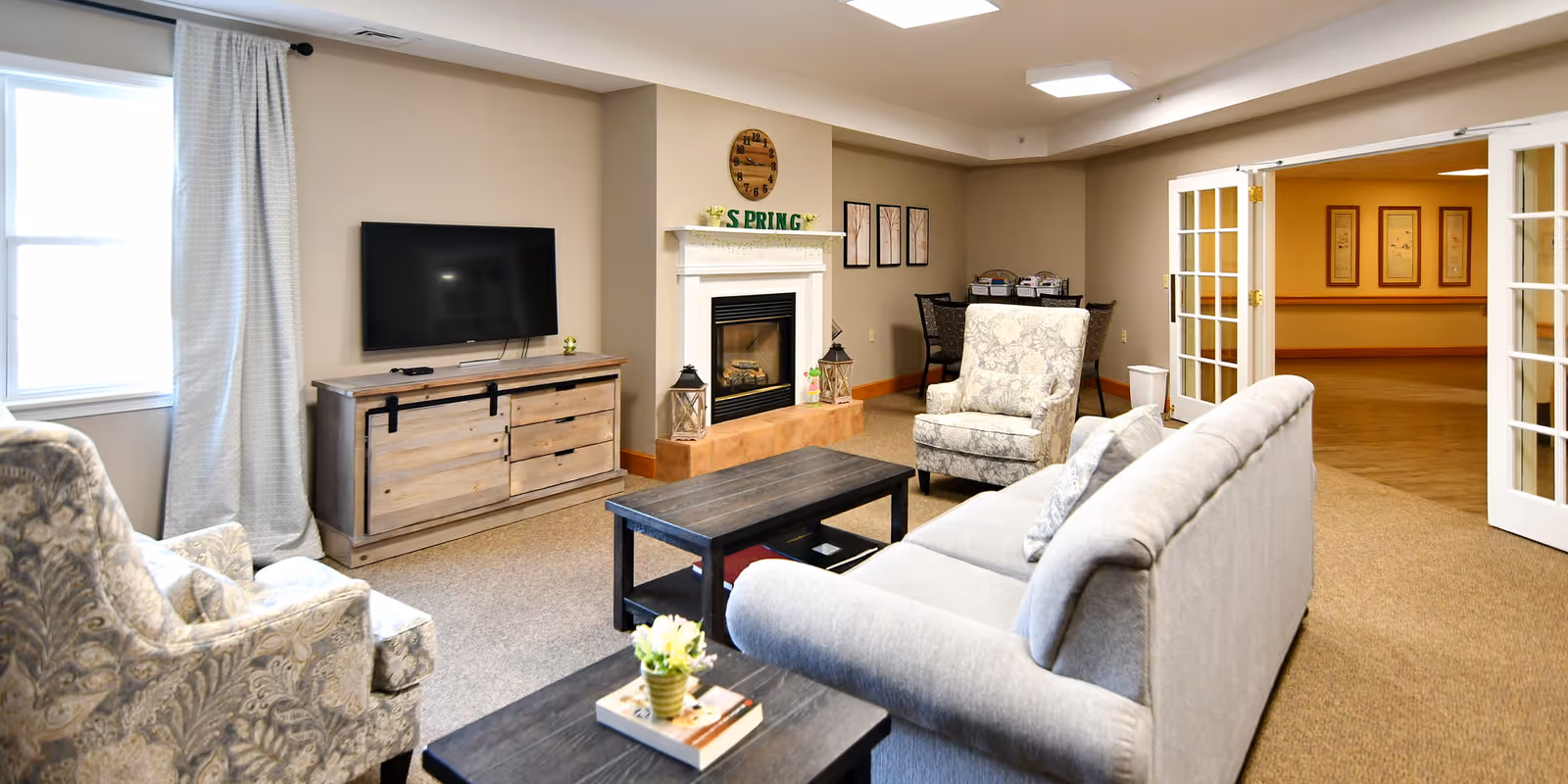 A cozy living room area with a gray sofa, two patterned armchairs, a wooden coffee table, and a TV mounted on the wall above a wooden cabinet. There is a fireplace with decorative items on the mantel, including a clock and a sign that says 'SPRING'. The room has beige walls, carpeted floor, and a window with light curtains. French doors lead to another room with framed artwork on the walls.