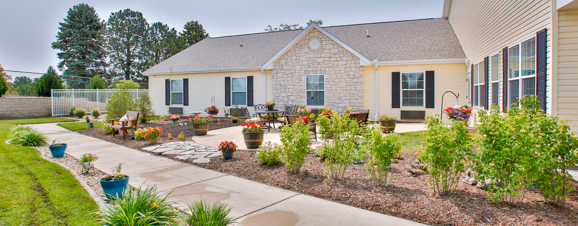 Outdoor patio area at Bickford of Bloomington featuring a paved walkway, garden beds with various plants and flowers, several potted plants, outdoor seating with chairs and tables, and a building with beige siding and stone accents in the background.