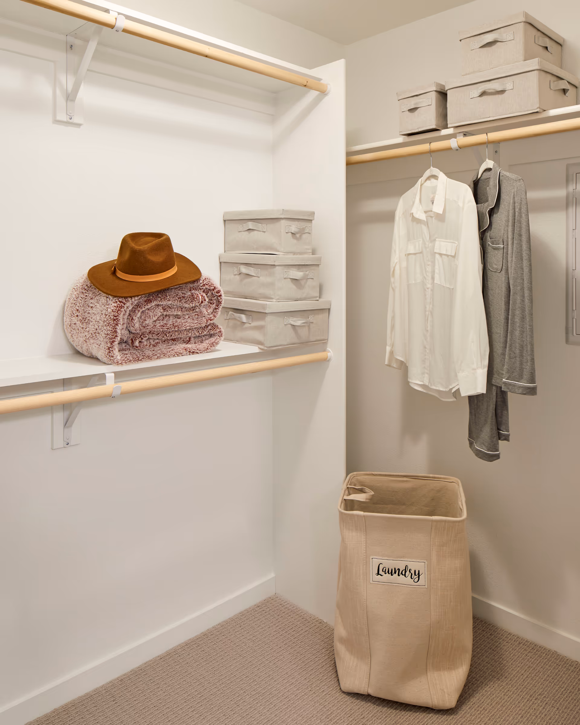 Neatly organized walk-in closet with hanging clothes, stacked storage boxes, a folded blanket topped by a hat, and a laundry hamper.