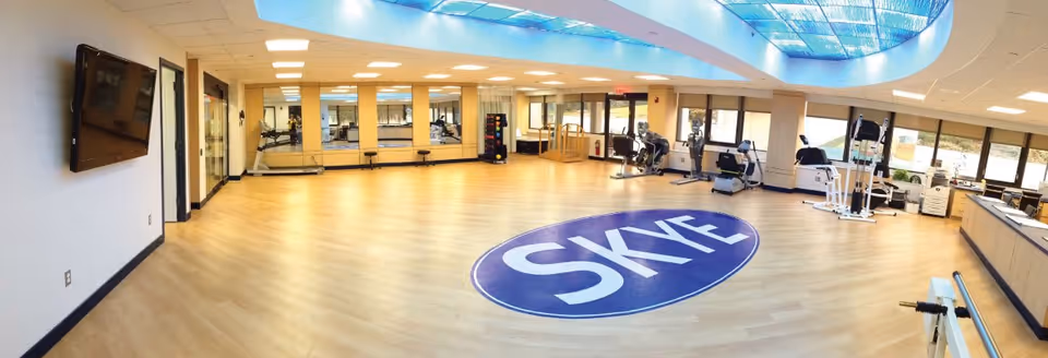 A spacious fitness room with light wood flooring and large windows allowing natural light. Exercise equipment such as stationary bikes and weight machines are arranged along the walls. The ceiling features a large skylight, and a blue circular logo with the word 'SKYE' is displayed prominently on the floor in the center of the room. A flat-screen TV is mounted on the left wall.