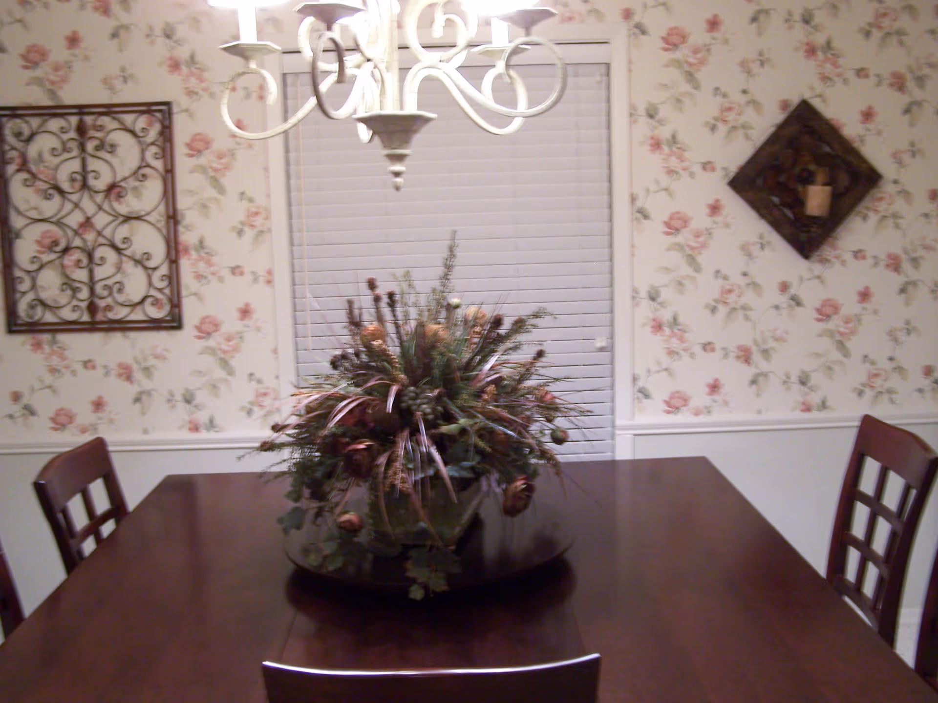 A dining room with a dark wooden table and four chairs. A large floral centerpiece sits in the middle of the table. The walls have floral wallpaper with a white wainscoting below. There is a window with closed white blinds behind the table, and decorative wall hangings on either side of the window. A white chandelier hangs above the table.