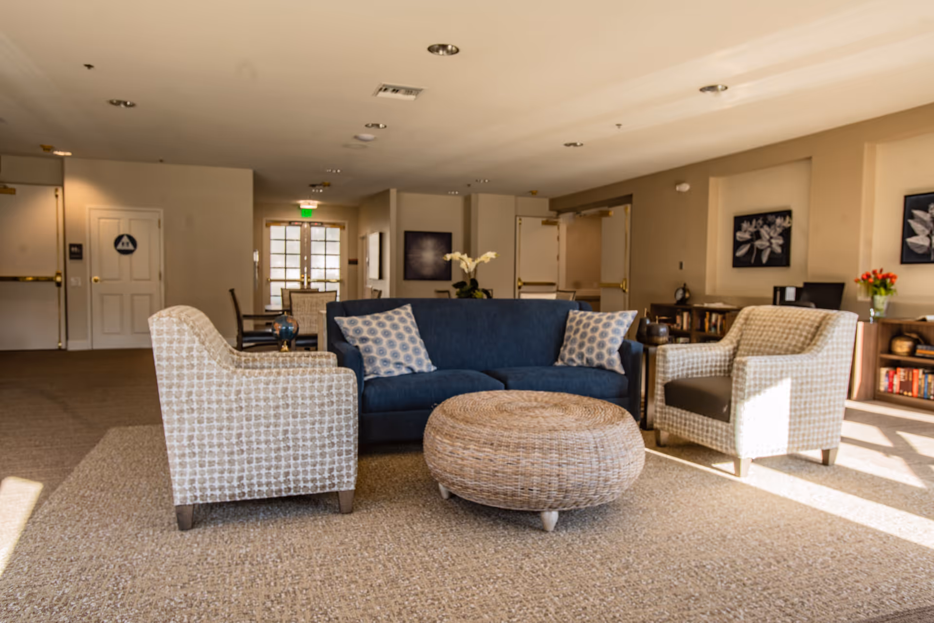 A cozy seating area in a senior living facility with a blue sofa, two patterned armchairs, and a round wicker ottoman in the center. The room has beige walls, carpeted floors, framed floral artwork on the walls, and a bookshelf with books and flowers. Sunlight streams in from windows on the right side.