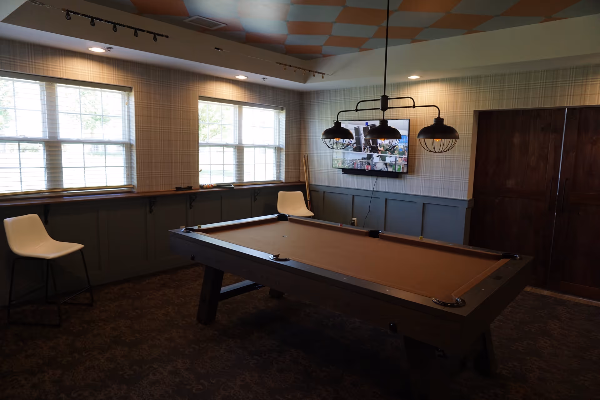 A room with a pool table in the center, two white chairs near the windows, a wall-mounted TV, and a ceiling light fixture with three lamps. The room has large windows with blinds, patterned wallpaper, and a carpeted floor.