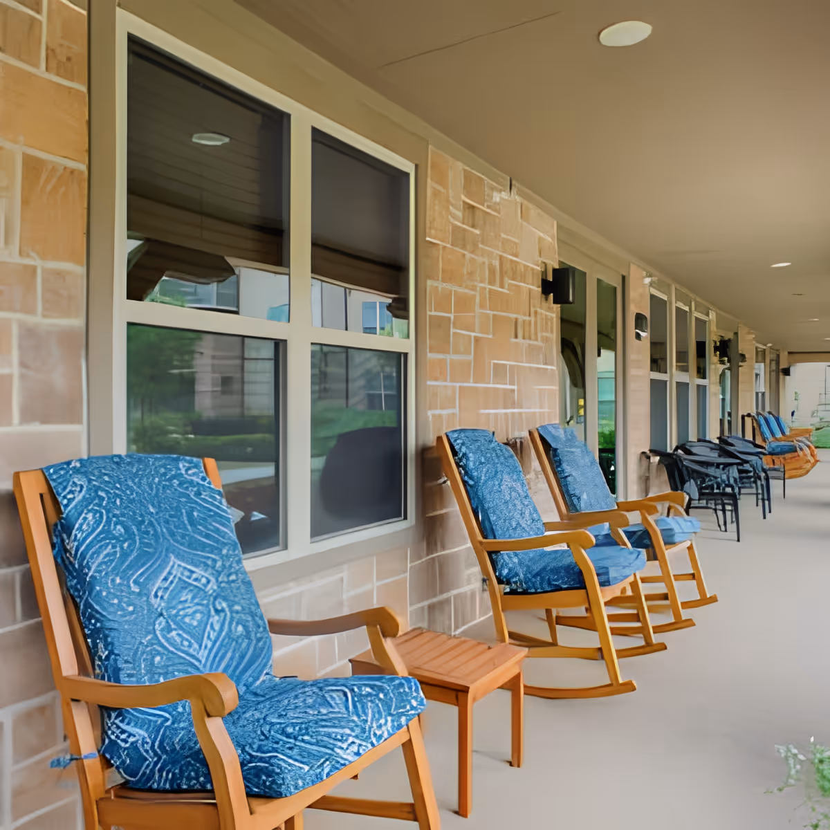 A covered outdoor patio area with a row of wooden rocking chairs featuring blue patterned cushions, small wooden side tables, and black metal chairs along a beige brick wall with large windows.
