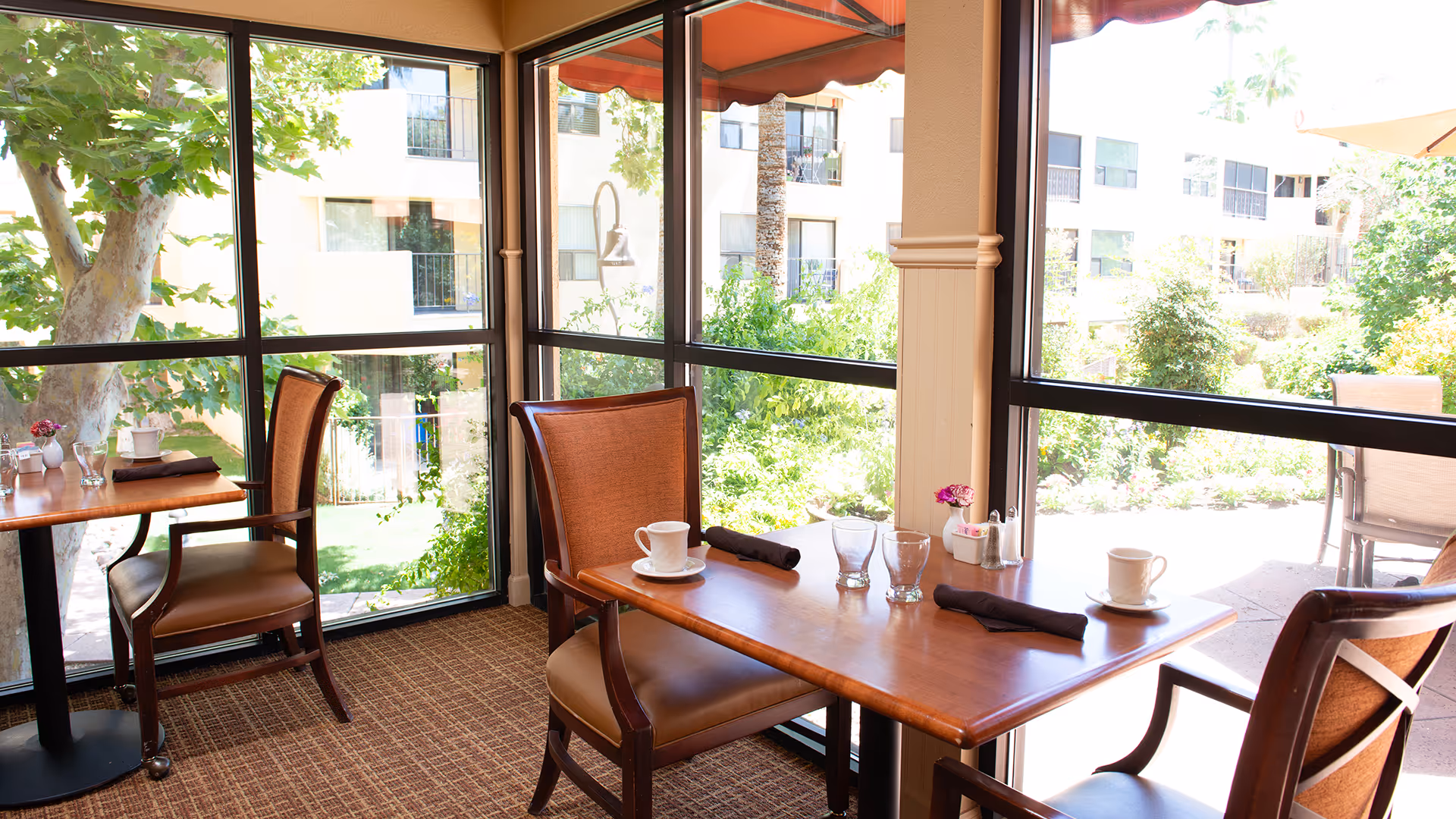 A bright dining area with wooden tables and cushioned chairs next to large windows overlooking a lush garden with trees and shrubs. The tables are set with cups, glasses, napkins, and small flower vases.