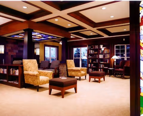 A warm wood-accented lounge/library with upholstered armchairs, ottomans, bookshelves and soft lighting.