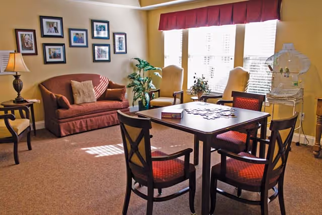 A cozy living room with a red sofa, two yellow armchairs near large windows with blinds, a wooden table with four chairs around it, a birdcage with two birds, a floor lamp, framed pictures on the wall, and a potted plant.