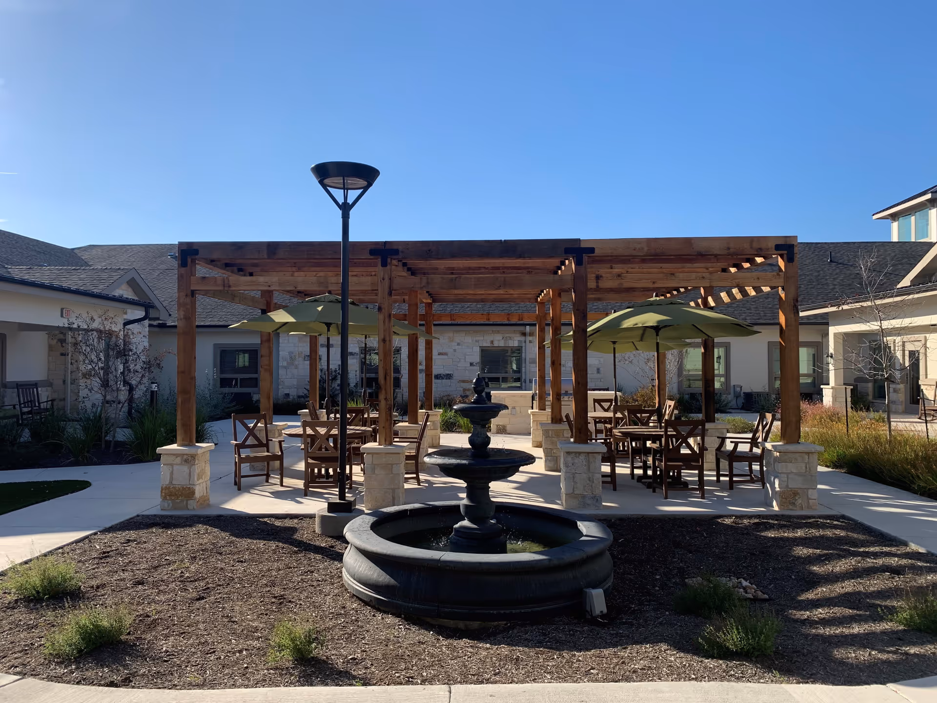 Outdoor courtyard area at Volante Senior Living of Sage Spring featuring a central black tiered water fountain, wooden pergolas with stone bases, multiple tables and chairs with green umbrellas, surrounded by a building under a clear blue sky.