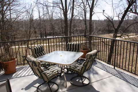 Outdoor patio area with a square glass table surrounded by four cushioned chairs with striped upholstery. The patio is enclosed by a metal railing and overlooks a wooded area with leafless trees and a dirt path in the background.