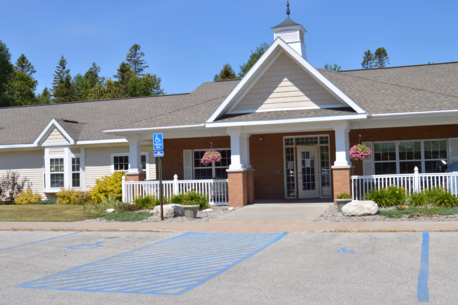 Front exterior view of Cedar Cove Assisted Living facility with a covered entrance supported by white columns, hanging flower baskets, and a handicap parking space in front. The building has a combination of brick and siding with a peaked roof and surrounded by greenery and trees.