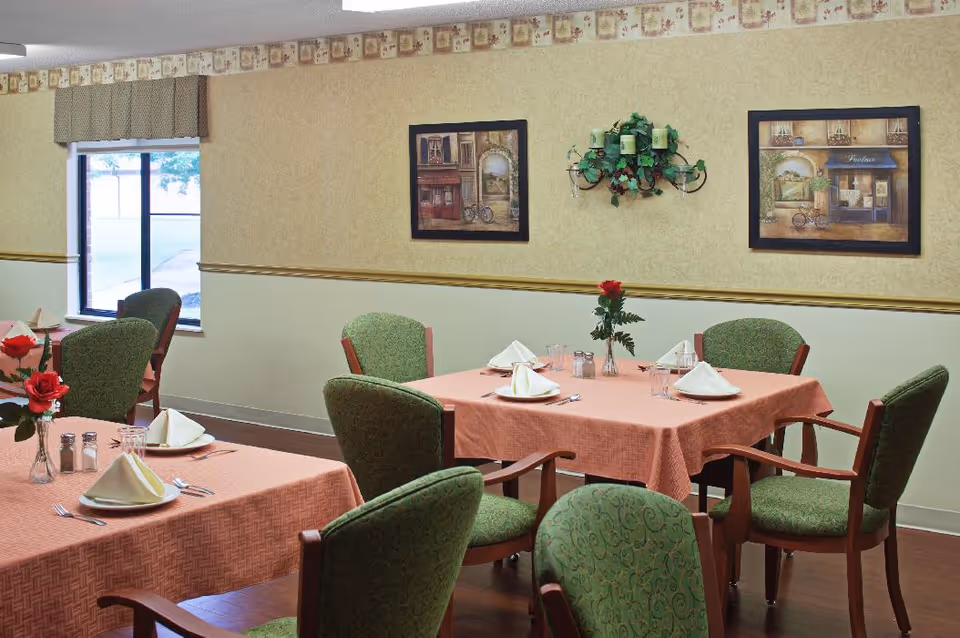A dining room with tables covered in peach-colored tablecloths, each set with white folded napkins, plates, glasses, and silverware. Green upholstered chairs surround the tables. The walls are decorated with framed paintings and a candle holder with greenery. A window with a valance lets in natural light.