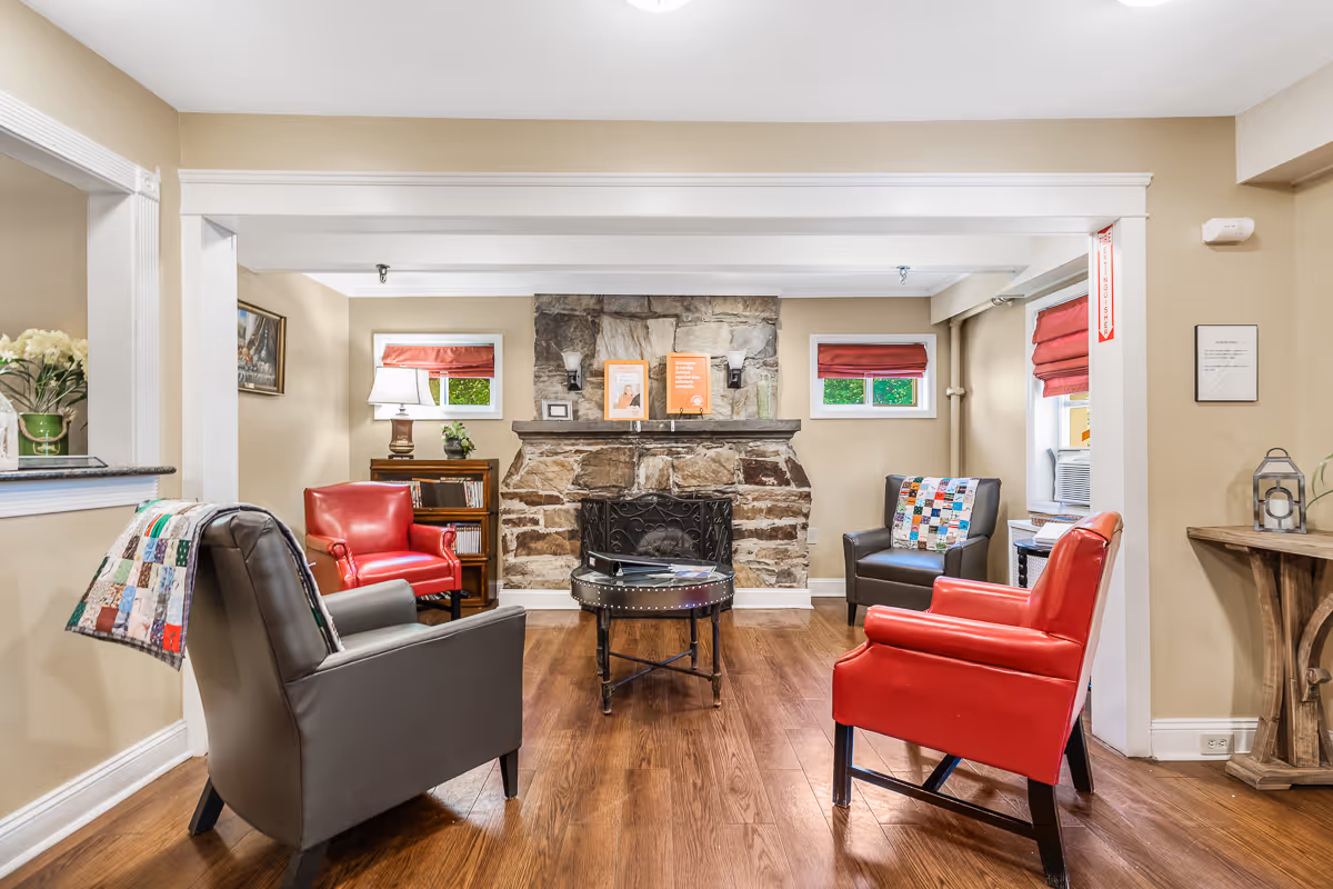 A cozy living room area with a stone fireplace in the center. The room features four armchairs arranged around a round coffee table, two red and two dark gray chairs, some with colorful patchwork quilts draped over them. There are two small windows with red blinds on either side of the fireplace, a wooden bookshelf with a lamp and plant on the left, and a small side table with a lantern on the right. The walls are painted beige and the floor is wooden.