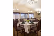 A dining room with multiple tables covered in white tablecloths, each set with green napkins and silverware. The room has several windows with curtains, wooden chairs with cushioned seats, and chandeliers hanging from the ceiling providing light.