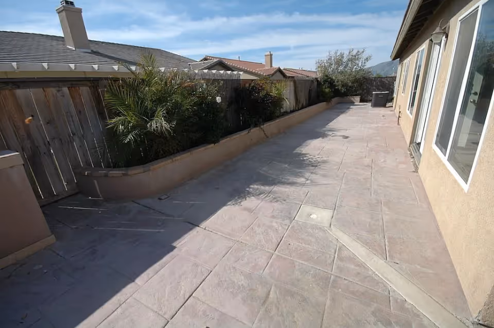 Long paved side patio with planter beds and a wooden fence beside a beige house under a blue sky.