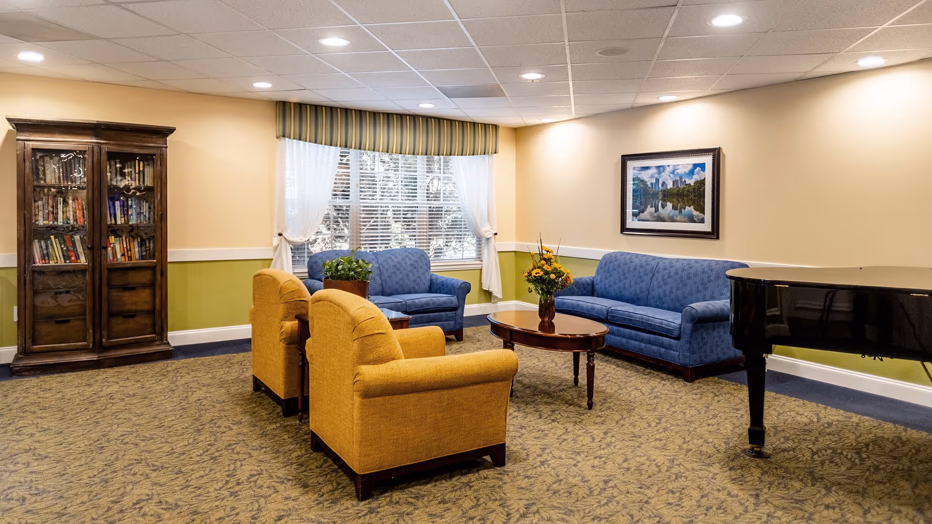 A cozy living room area with two blue sofas, two yellow armchairs, a wooden coffee table with a flower vase, a large window with white curtains and a striped valance, a wooden cabinet filled with books, a framed picture on the wall, and a black grand piano on the right side.