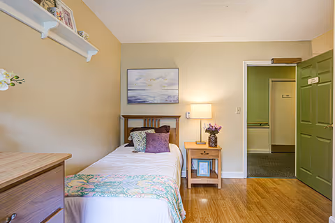 A small, neatly arranged bedroom with a single bed against the wall. The bed has white bedding with a colorful quilt at the foot and several decorative pillows. Next to the bed is a wooden nightstand with a lamp and a vase of flowers. Above the bed hangs a framed painting of a seascape. The room has light-colored walls, wooden flooring, and an open green door leading to a hallway.