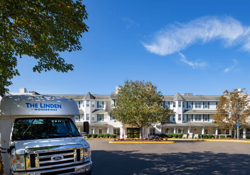 Front exterior view of a senior living facility with a white multi-story building, large windows, and a covered entrance. A white shuttle bus with the text 'THE LINDEN AT WOODBRIDGE' is parked in front. Trees and a clear blue sky are visible.