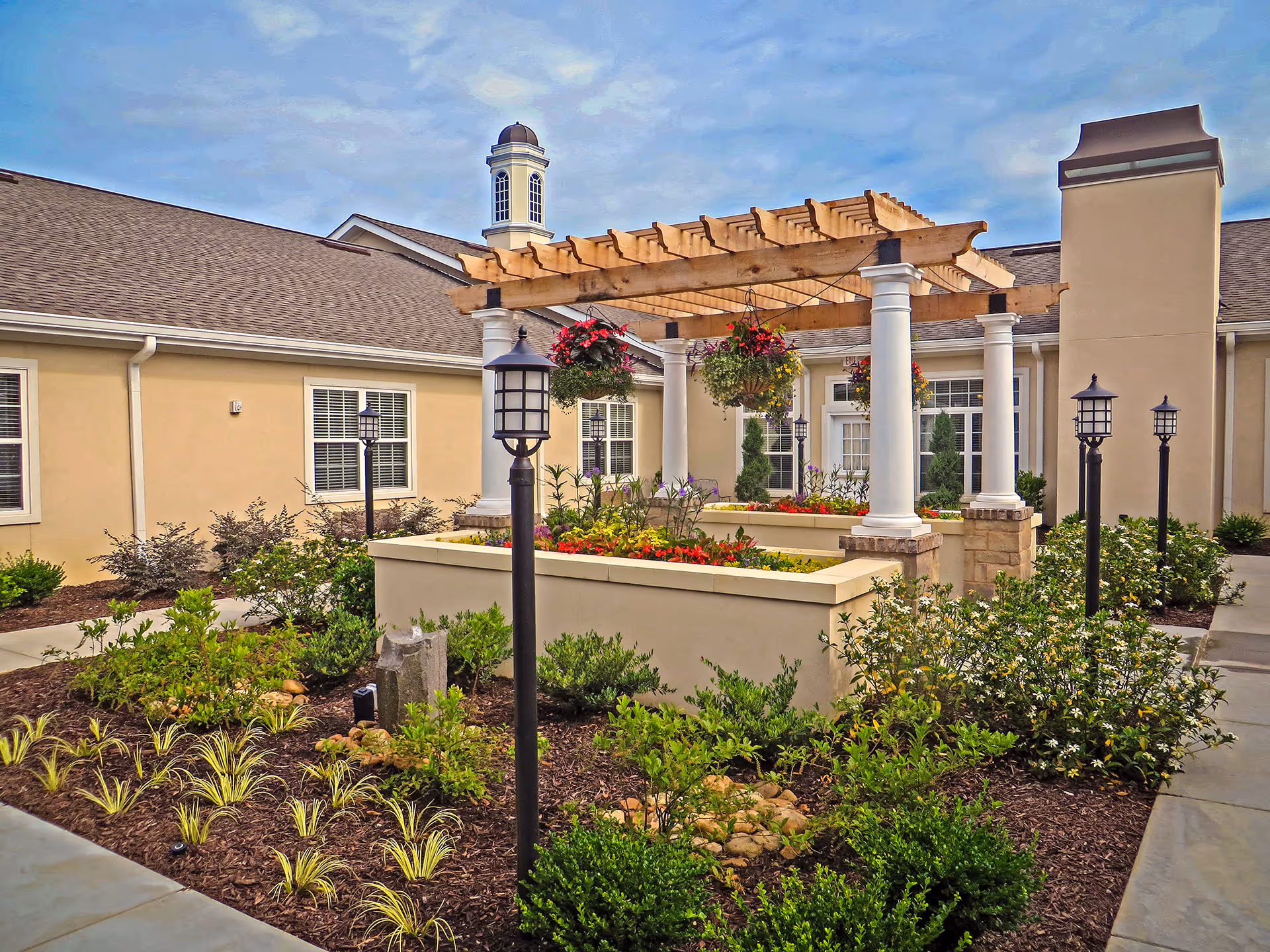 Outdoor courtyard area at Benton Manor Of Stockbridge featuring a wooden pergola with hanging flower baskets, surrounded by landscaped garden beds with various plants and shrubs. The building exterior is beige with white trim, and there are multiple black lamp posts lining the paved walkways.