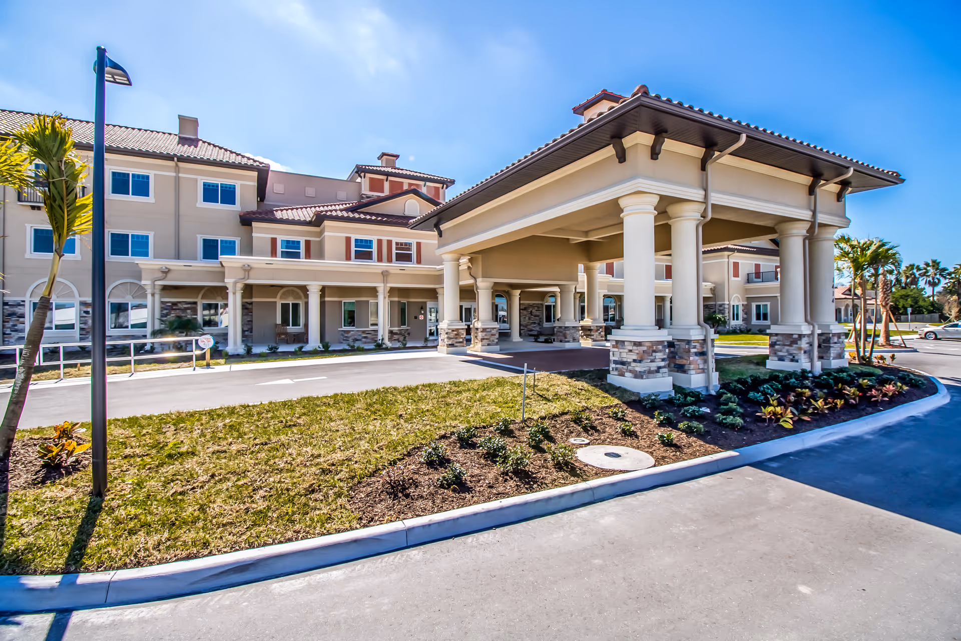 Front entrance of a multi-story senior living building with a covered porte-cochere, columns, landscaped driveway, and palm trees.