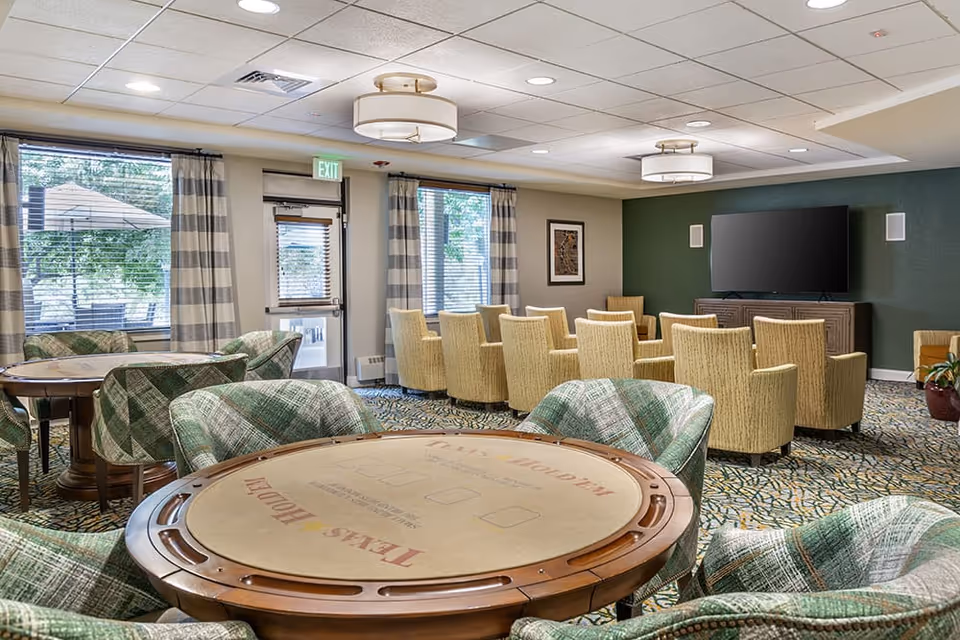 A spacious common area with multiple round tables and cushioned chairs arranged for group activities or socializing. The room features large windows with striped curtains, a green accent wall with a mounted flat-screen TV, and beige armchairs facing the TV. The carpet has a patterned design, and ceiling lights provide ample illumination.