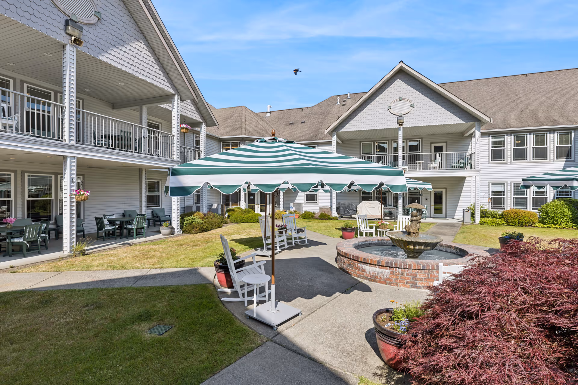 Outdoor courtyard area of a senior living facility with a central brick fountain, white rocking chairs, and green and white striped umbrellas. The courtyard is surrounded by a two-story building with balconies and patio seating. There are potted plants and well-maintained landscaping under a clear blue sky.
