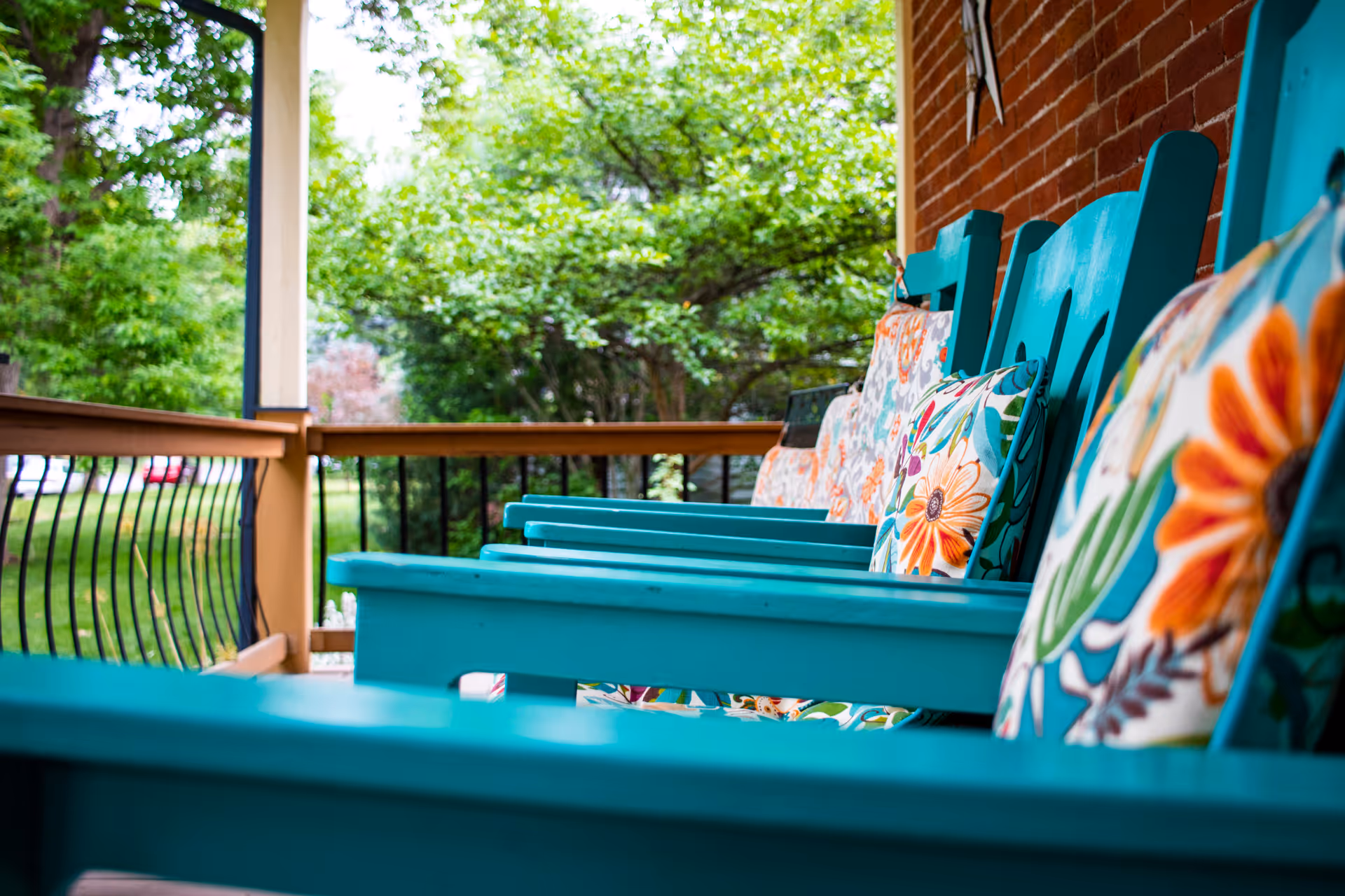 Close-up view of a porch with turquoise wooden chairs lined up, each with colorful floral cushions. The porch has a wooden railing with black metal balusters and a brick wall on one side. Green trees and grass are visible in the background.