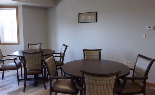 Small communal dining area with round wooden tables, upholstered chairs, and a wall sign reading "SWEET".