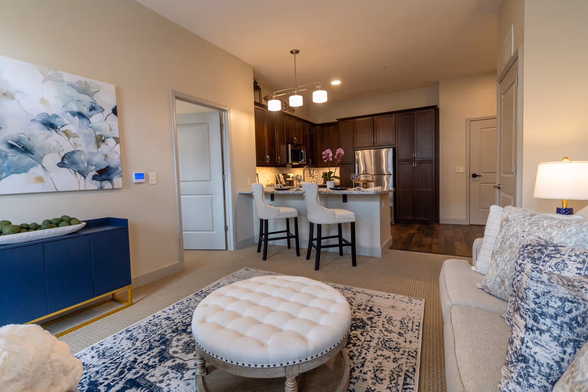 Open living area showing a tufted round ottoman, a sofa with patterned pillows, and a kitchen island with two white bar stools and dark wood cabinets.