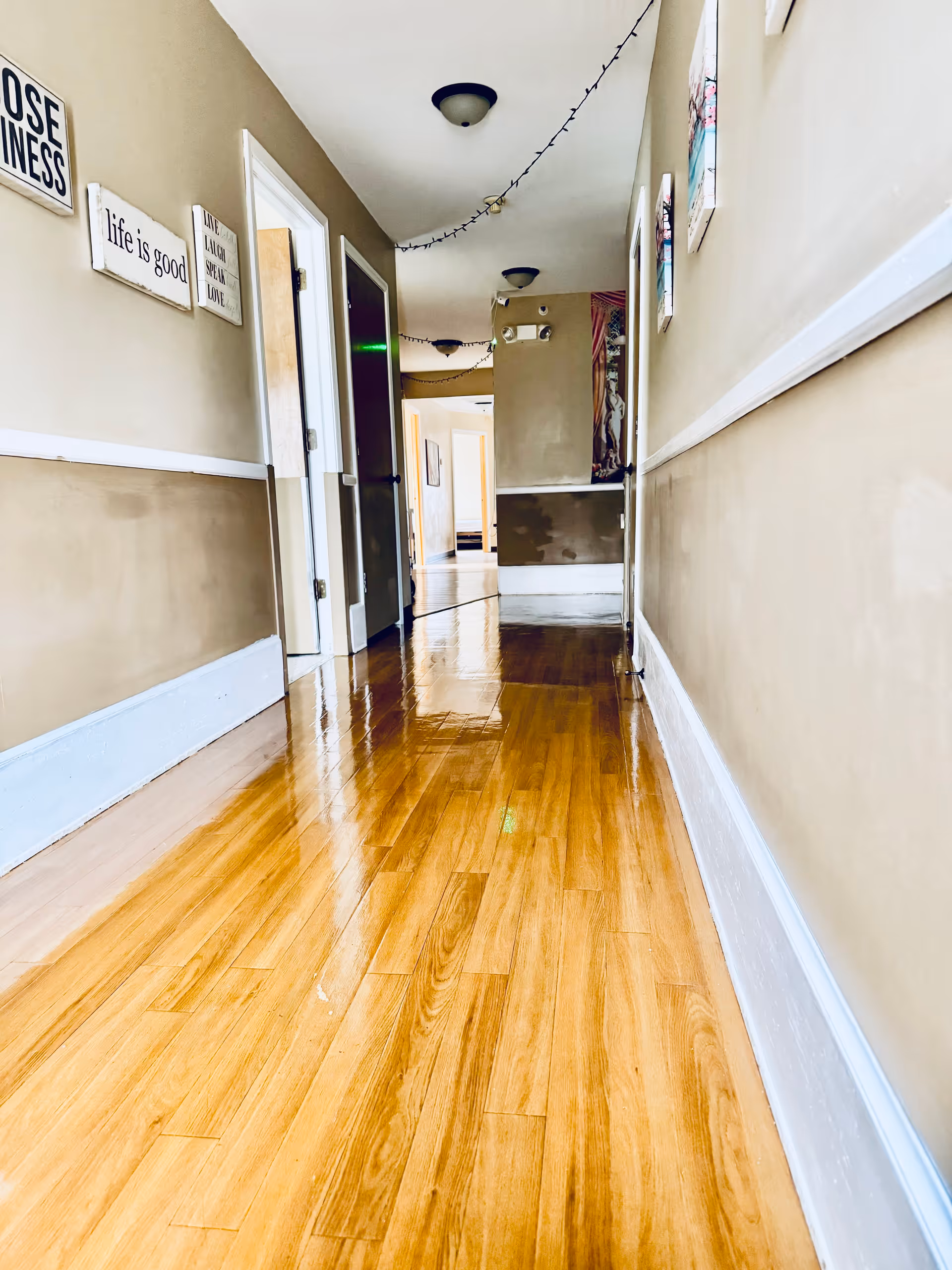 A long, narrow hallway with polished wooden floors and beige walls. Several doors line the hallway, and there are framed inspirational quotes on the left wall. Ceiling lights and string lights are visible overhead.