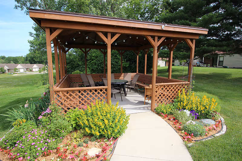 Wooden gazebo with chairs and a table, surrounded by flower beds and a paved walkway on a grassy lawn.
