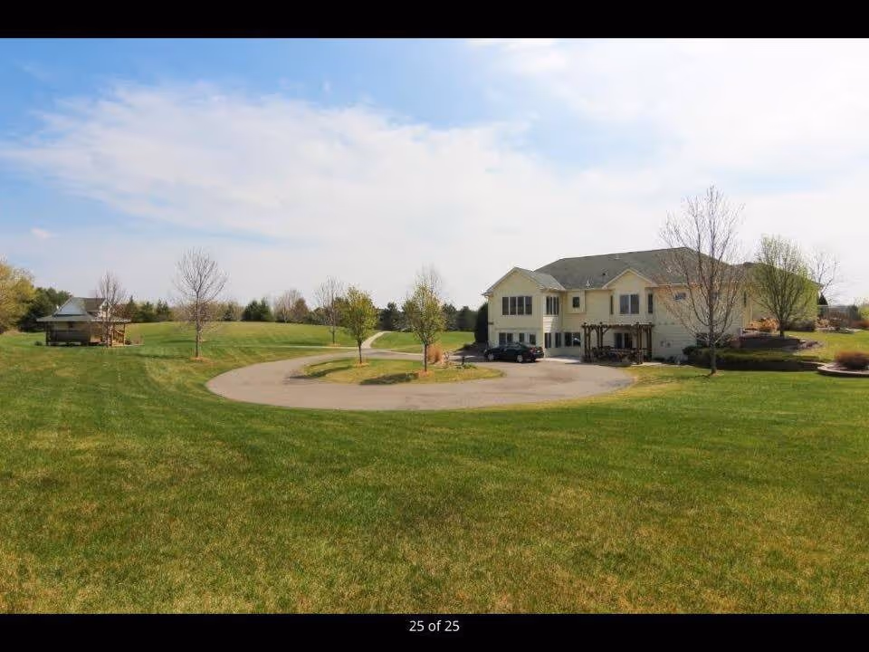 A large two-story beige building with a gray roof situated in a spacious green lawn area with a circular driveway. There are several leafless and leafy trees scattered around the lawn, and a small gazebo structure is visible on the left side. The sky is partly cloudy with blue patches.