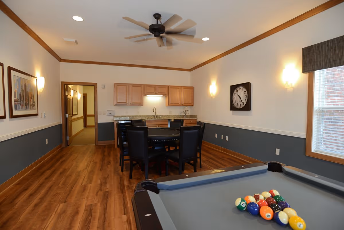Communal interior room with a pool table in the foreground, a dining table and kitchenette in the background.