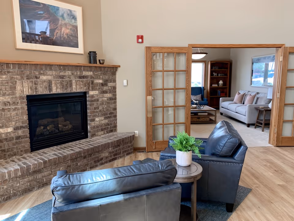 Common living area with a brick fireplace, two leather chairs around a small table with a potted plant, and a view into an adjacent sitting room with sofas.