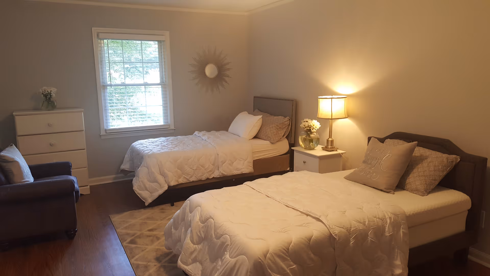 A cozy bedroom with two single beds, each with white bedding and pillows. Between the beds is a small white nightstand with a lamp and a vase of flowers. There is a window with white blinds on the left wall, a white dresser with a small flower vase on top, and a dark armchair next to it. The walls are painted a light beige color, and a decorative sunburst mirror hangs above the bed near the window. The floor is wooden with a patterned area rug partially visible.