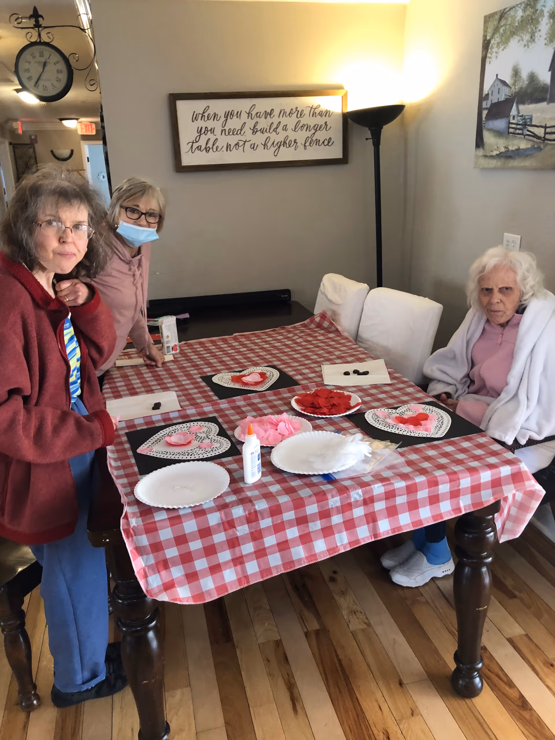Three elderly women are gathered around a table covered with a red and white checkered tablecloth. The table has craft supplies including paper hearts, glue, and plates. One woman is standing on the left, another is standing in the middle wearing a face mask, and the third woman is seated on the right wrapped in a white blanket. The room has wooden floors, a standing lamp, and wall decorations including a clock and framed signs.