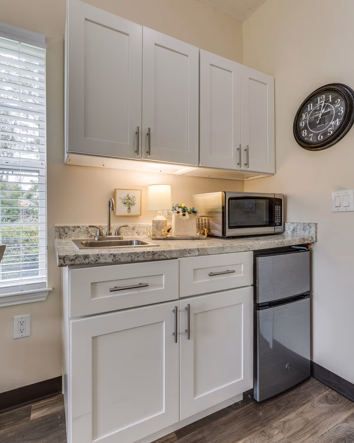 Small kitchen area with white cabinets, a granite countertop, a stainless steel sink, a small lamp, a basket with flowers, a microwave, and a compact refrigerator. A window with blinds is on the left, and a round black wall clock is on the right wall.