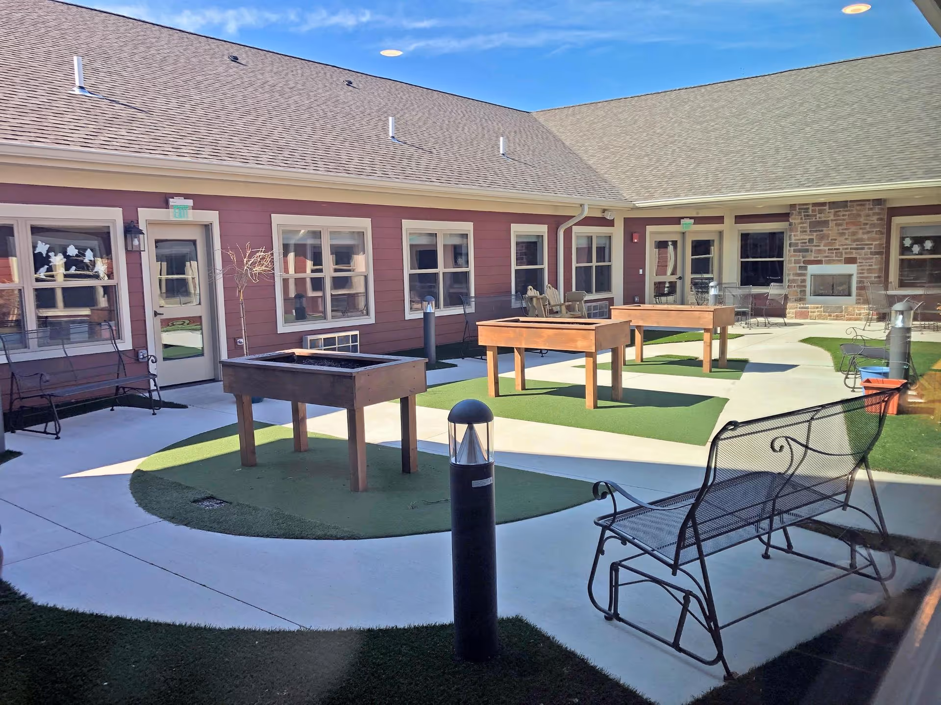 Outdoor courtyard area of a retirement community with several wooden game tables on green artificial turf, metal benches, and a stone fireplace attached to the building. The building has red siding and multiple windows and doors leading to the courtyard.