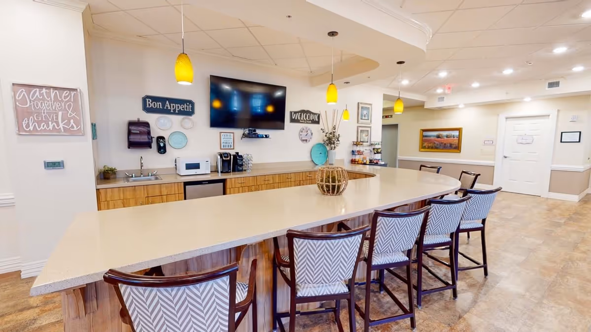 A bright and welcoming common area with a large curved countertop surrounded by six patterned chairs. The countertop has a decorative basket centerpiece. Behind the counter is a kitchenette with a sink, microwave, coffee maker, and various wall decorations including signs that say 'Bon Appetit' and 'Welcome'. The floor is tiled, and the ceiling has recessed lighting and hanging yellow pendant lights.