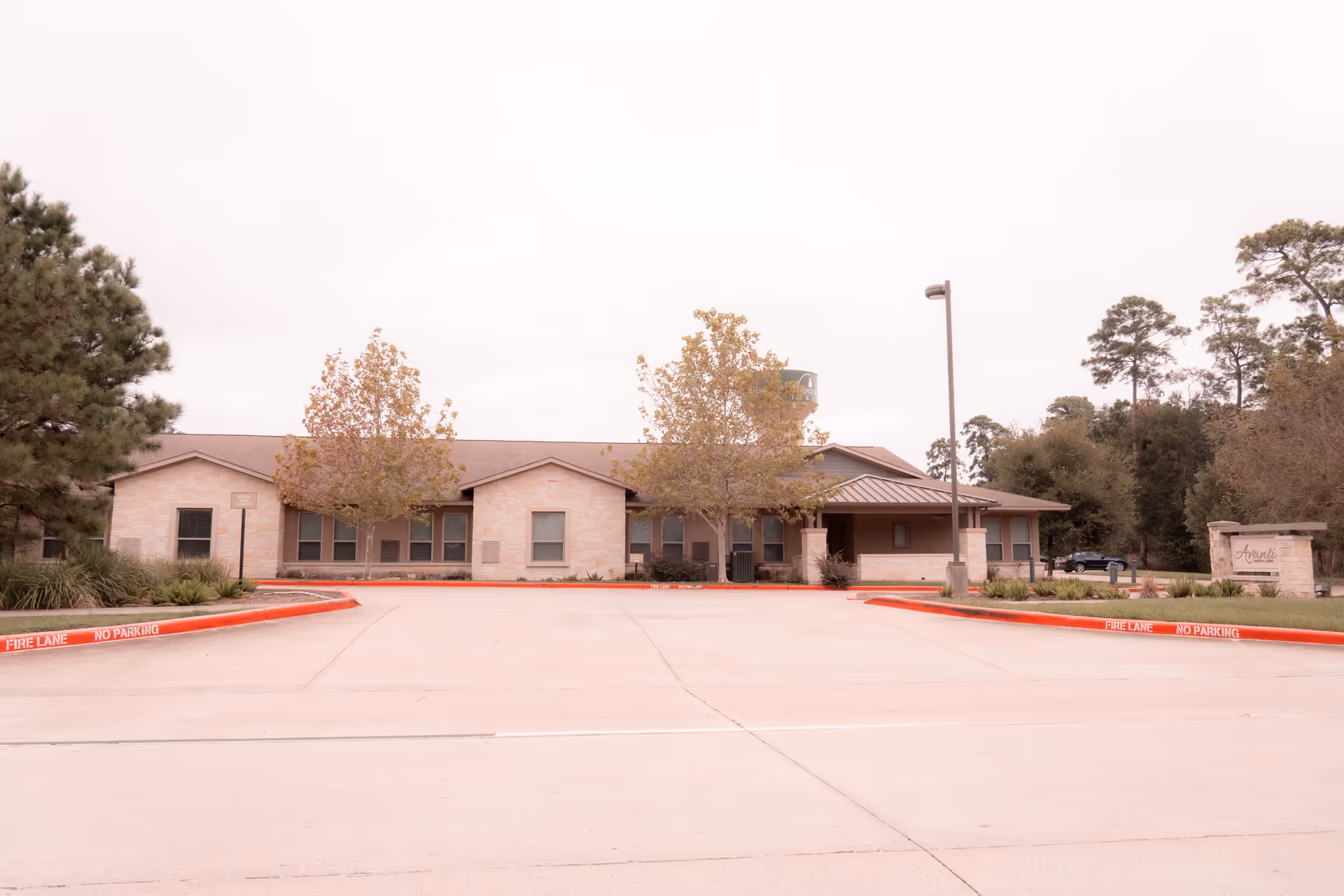 Exterior view of a single-story senior living facility building with a stone facade, several windows, and a covered entrance. There are trees and landscaping around the building, a water tower visible in the background, and a parking area with red curbs marked 'FIRE LANE NO PARKING'. A sign near the entrance reads 'Avanti Senior Living at Vision Park'.