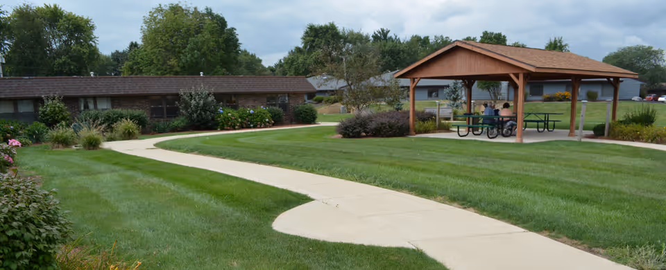 A well-maintained outdoor area at Valparaiso Care and Rehabilitation featuring a curved concrete walkway through green grass, a wooden gazebo with picnic tables underneath, and a few people sitting inside the gazebo. Surrounding the area are bushes, trees, and a single-story building in the background under a cloudy sky.